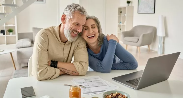 A couple in their middle age, both with grey hair, leaning affectionately towards each other with smiles, engaged in reviewing documents and various items spread out on a computer in a well-lit and stylishly minimalistic room.