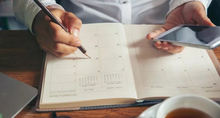 A person wearing a button-up shirt sits at a table, diligently writing in a planner book with a pencil. A cup of tea sits nearby, adding to the serene ambiance of the organized planning session.