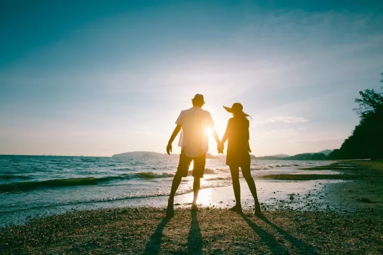 A romantic scene unfolds on a beach at sunset, as a woman and a man hold hands, their silhouettes against the backdrop of a colorful sky.