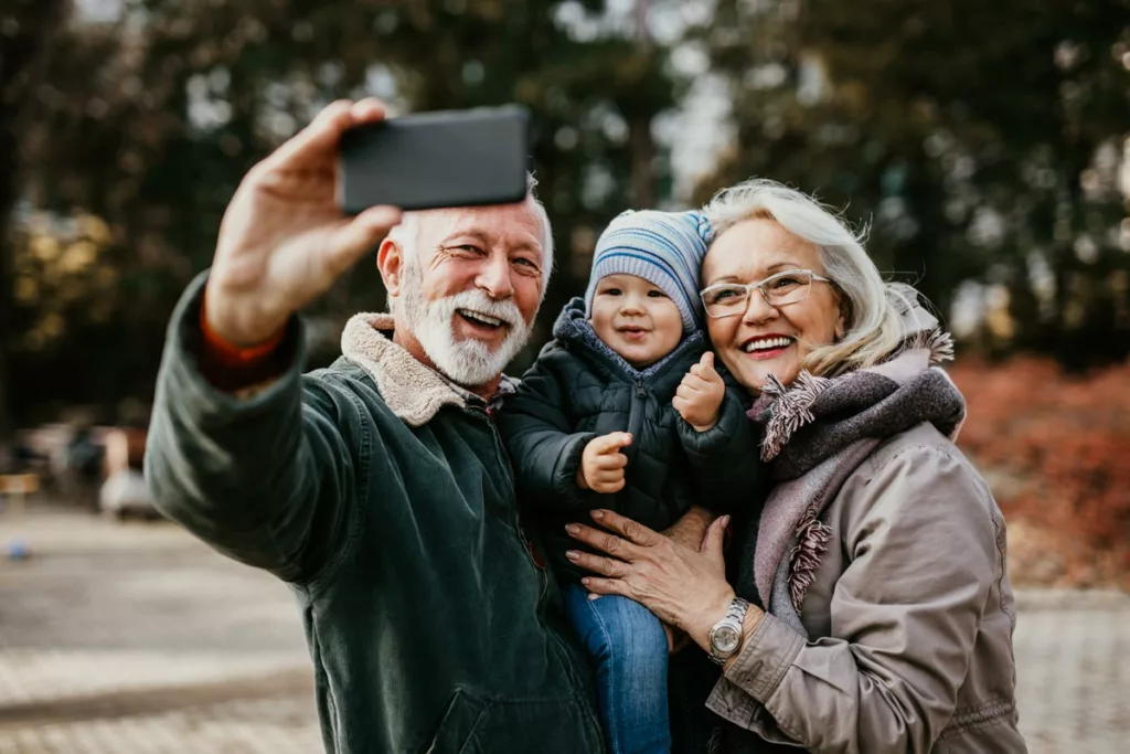 An older couple joyfully embraces while a baby holds a smartphone, capturing a selfie of the three of them.